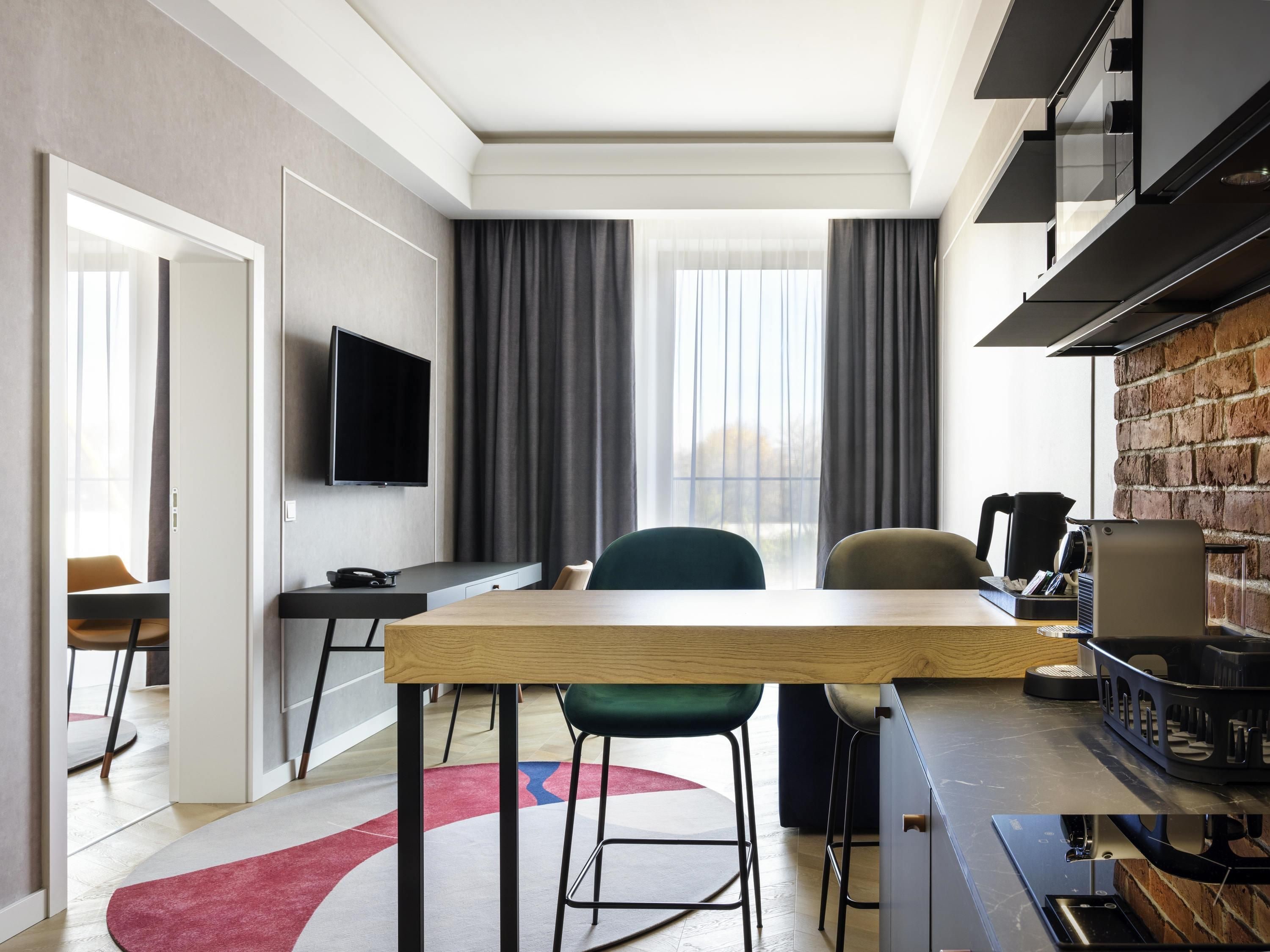 A modern hotel room with a dining table, chairs, a TV, a coffee machine, and a brick wall.