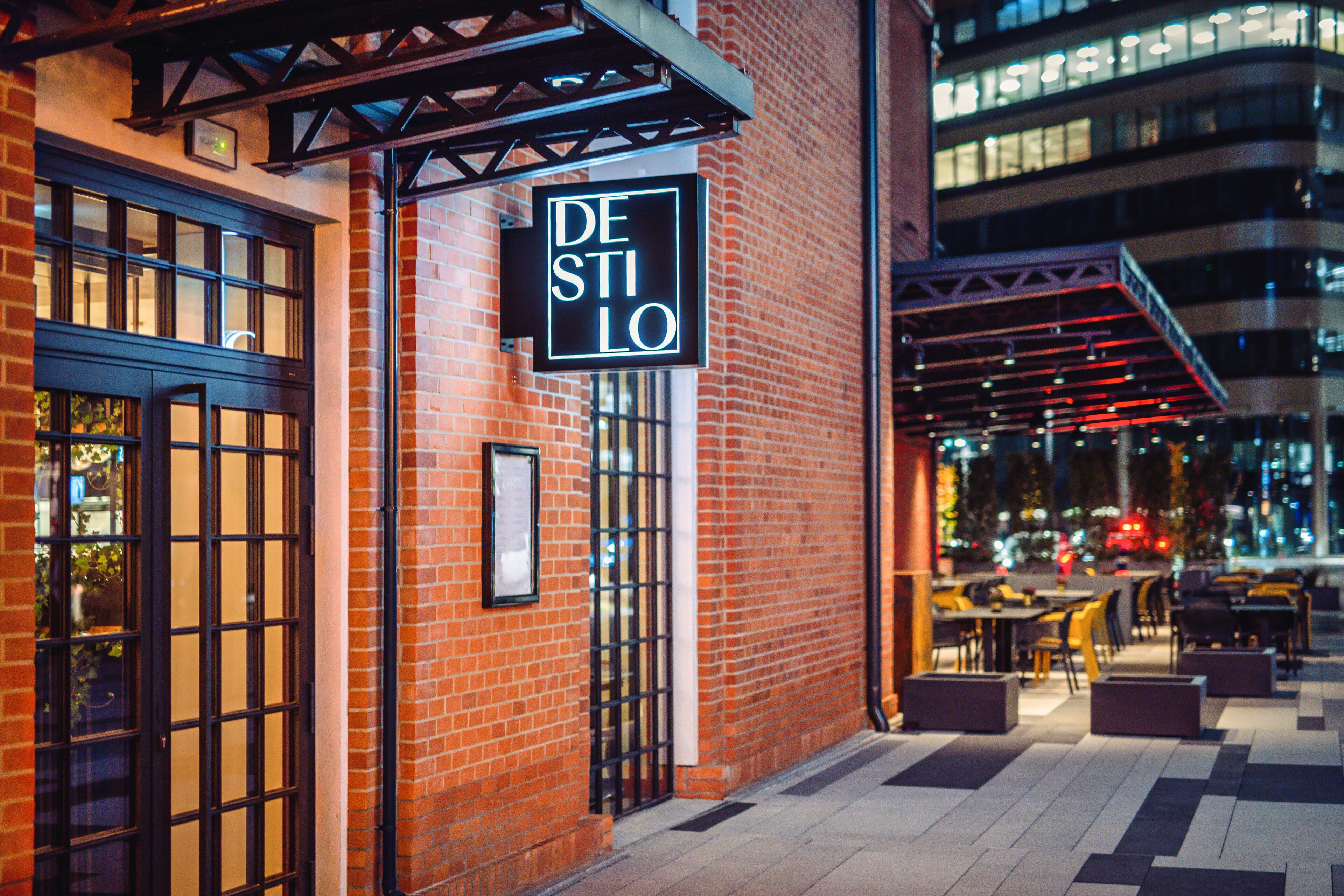 A brick building with a sign reading "DE STILO" and outdoor seating area with tables and chairs.