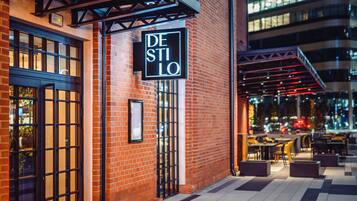 A brick building with a sign reading "DE STILO" and outdoor seating area with tables and chairs.