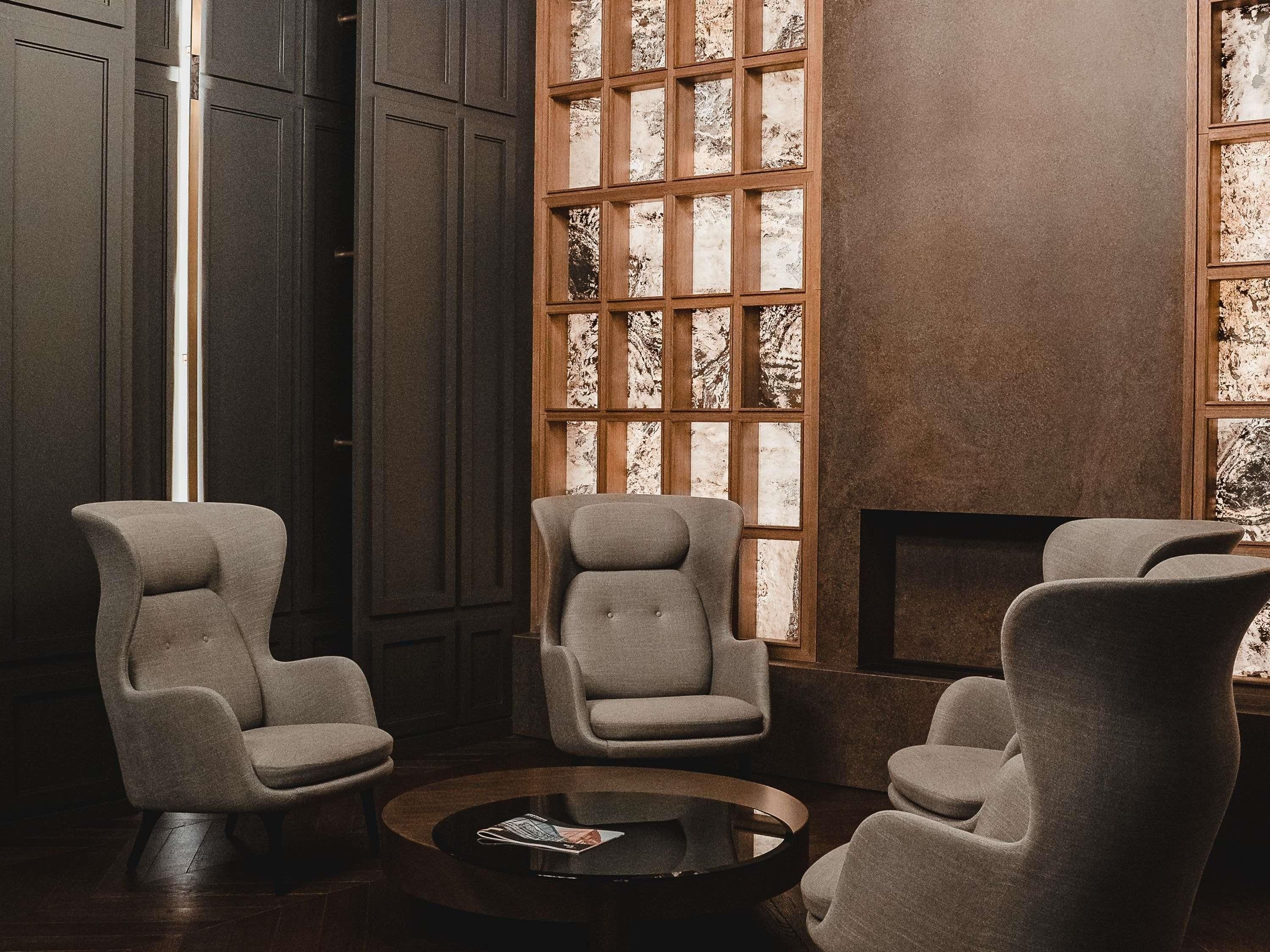 Ein moderner Lounge-Bereich mit drei grauen Sesseln, einem runden Couchtisch und einem großen Fenster mit Holzgitter-Design.