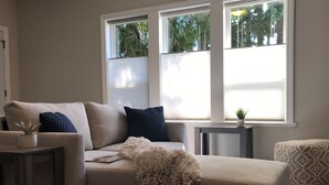 Ett vardagsrum med en beige soffa, en mjuk sittpuff, ett litet bord med en växt och en blå, mönstrad matta.