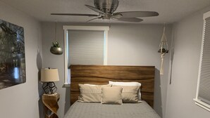Une chambre à coucher comprenant un lit, un ventilateur de plafond, une fenêtre avec des stores, une lampe fixée au mur et un tapis.