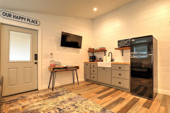 A kitchen with a flat-screen TV, a Galanz refrigerator, wooden cabinets, and a sign that reads 'OUR HAPPY PLACE'. - Barnside Hangout - Ranch Retreat - Master Spa - Hot Tub - Near Fredericksburg (Fredericksburg)