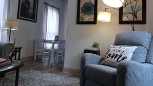 Ein Wohnzimmer mit blauem Ohrensessel, einem Esstisch mit Stühlen, einer Lampe und gerahmten Kunstwerken an den Wänden.