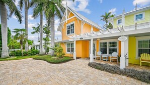 A two-story building with a yellow and white exterior, a covered patio with chairs, and a paved driveway. - Cozy Retro Cottage located less then 2 blocks from the Gulf Beach - Heated Pool in Private Oasis! (Holmes Beach)