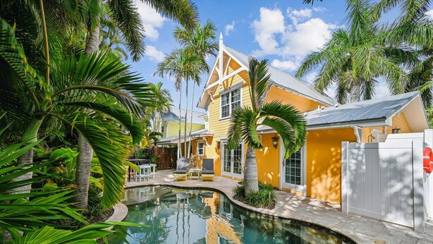 A pool area with a curved edge, surrounded by palm trees and a yellow building with a triangular roof. - Cozy Retro Cottage located less then 2 blocks from the Gulf Beach - Heated Pool in Private Oasis! (Holmes Beach)