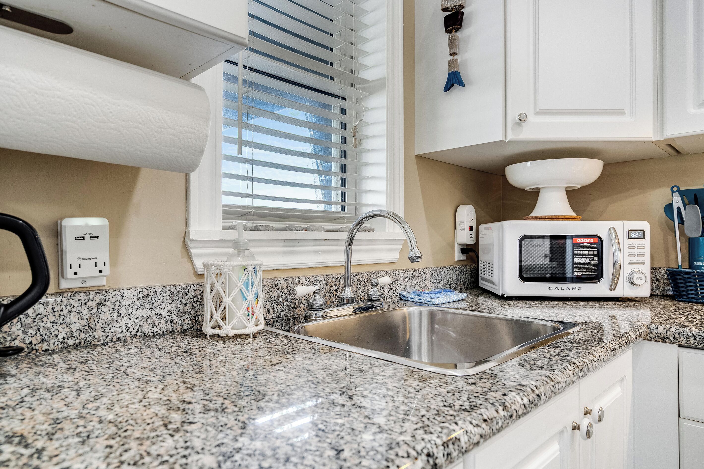 A kitchen with a granite countertop, stainless steel sink, white cabinets, a microwave, and a window with blinds.