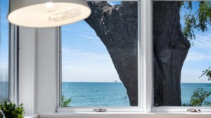 A room with a table and a chair, a lamp, and a view of the sea through a window with a tree in the foreground.