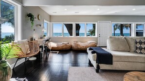 A spacious living room with a large window overlooking the sea, a cozy seating area with a sofa and bean bags, and a dining area with a table and chairs.