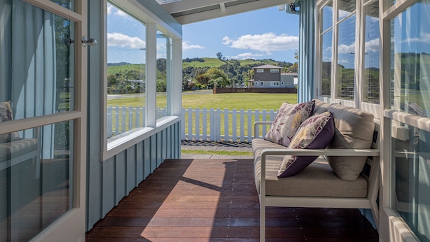 Un porche cubierto con terraza de madera, un banco blanco con cojines y vista a un paisaje verde y una casa a lo lejos.
