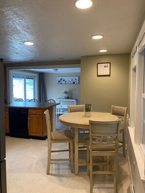 A dining area with a round table and six chairs, a kitchen with a dishwasher, and a window with curtains.