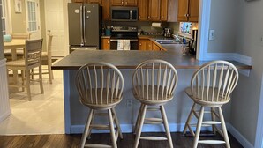 Een keuken met houten kastjes, een roestvrijstalen koelkast, een magnetron en een spoelbak. Er staan drie houten krukken bij het keukeneiland.