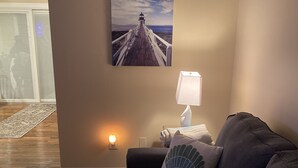 Een woonkamer met houten vloer, een grijze bank met decoratief kussen, een aan de muur bevestigd beeld van een pier en een wandlamp.