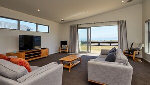 A modern living room with a flat-screen TV, grey sofas, a wooden coffee table, and a large sliding glass door leading to a balcony with a view of the beach. - The Kaikoura Bach Retreat (Kaikoura)