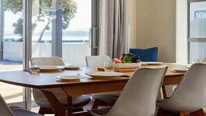 A dining area with a table set for four, a bowl of fruit, and a view of the sea through large windows. - The Kaikoura Bach Retreat (Kaikoura)