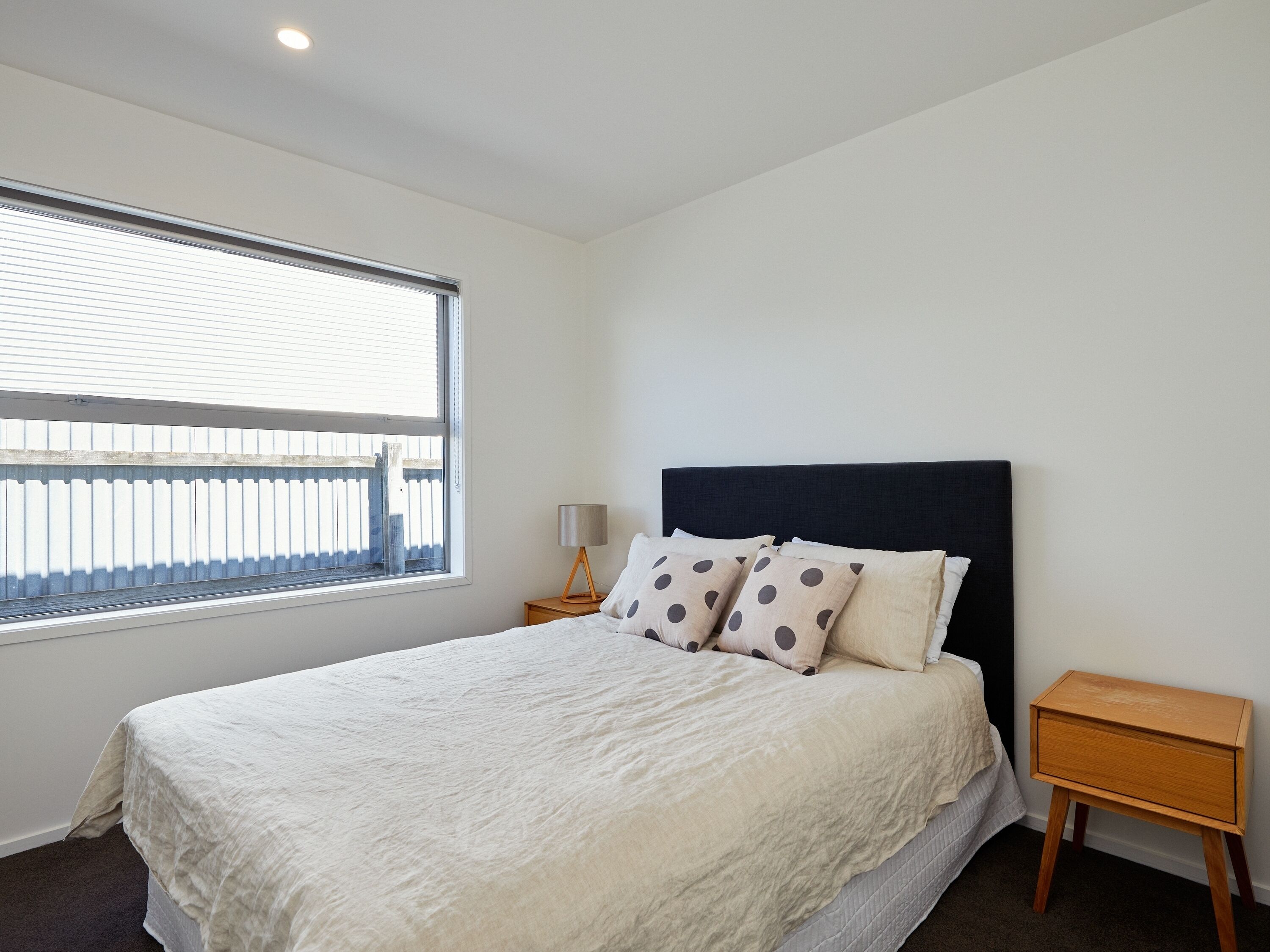A bedroom with a large window, a bed with white bedding, a dark headboard, and a wooden nightstand with a lamp.