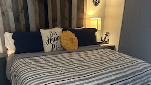 Un lit bien fait, avec une literie à rayures, un coussin décoratif orné d’un motif, et une table de chevet fixée au mur, sur laquelle se trouvent une lampe et une décoration en forme d’ancre.