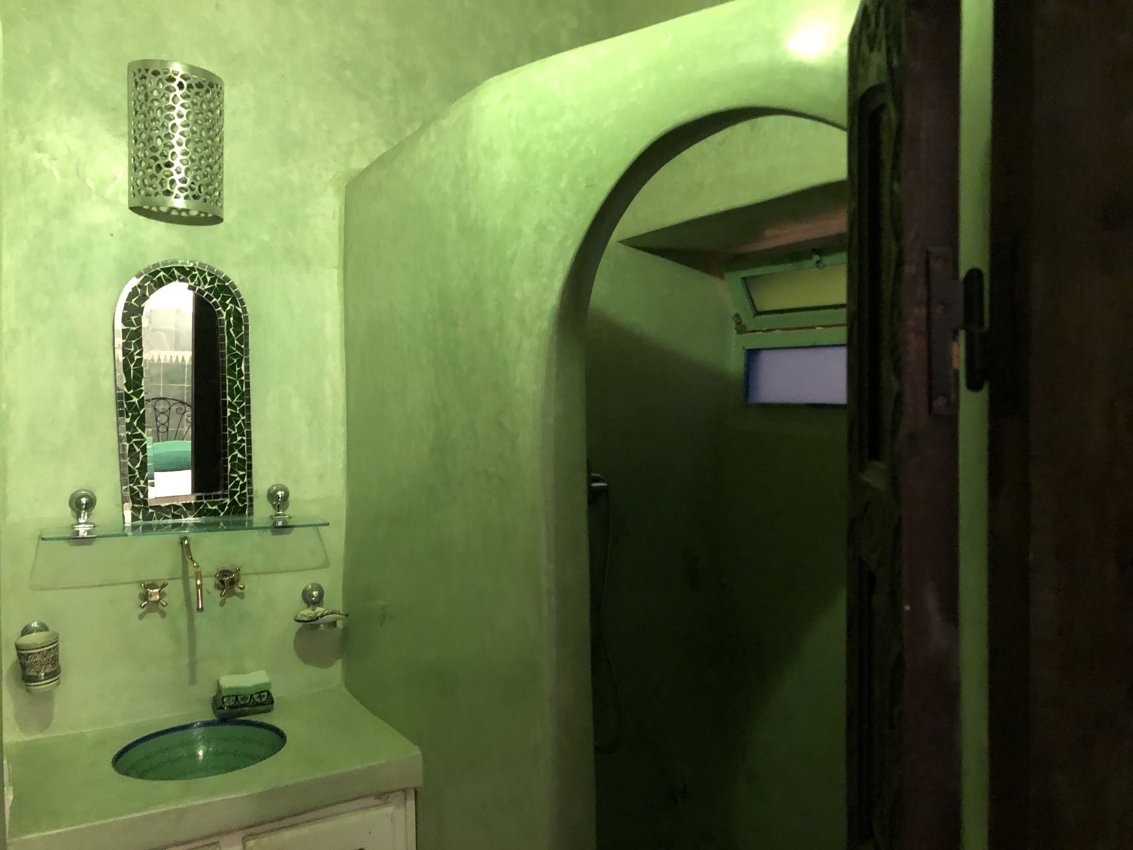 Ein Badezimmer mit grüner Wand, einem Glaswaschbecken, einem Spiegel mit Mosaikrahmen und einer kleinen Ablage für Toilettenartikel.