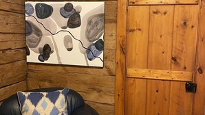 A log cabin interior with a wooden door, a leather armchair with a patterned pillow, and a painting of abstract shapes on the wall.