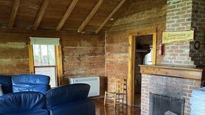 A rustic cabin interior with a wooden ceiling, a brick fireplace, a blue leather sofa, and a wooden table with a flower vase.