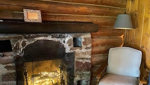 Interior rústico de una cabaña con chimenea de piedra, una silla de madera y una lámpara.