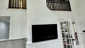A spacious living area with a high ceiling, a fireplace, a television, and a staircase with a railing.