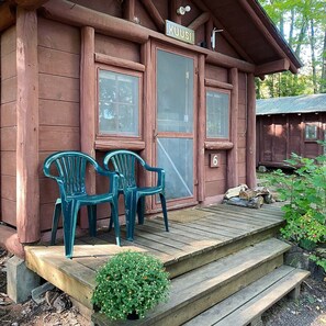 A rustic cabin with a wooden porch, two plastic chairs, and a small potted plant. - Kuusi (#6) - Cabin on Otter Lake, Manninen's Cabins (Chassell)