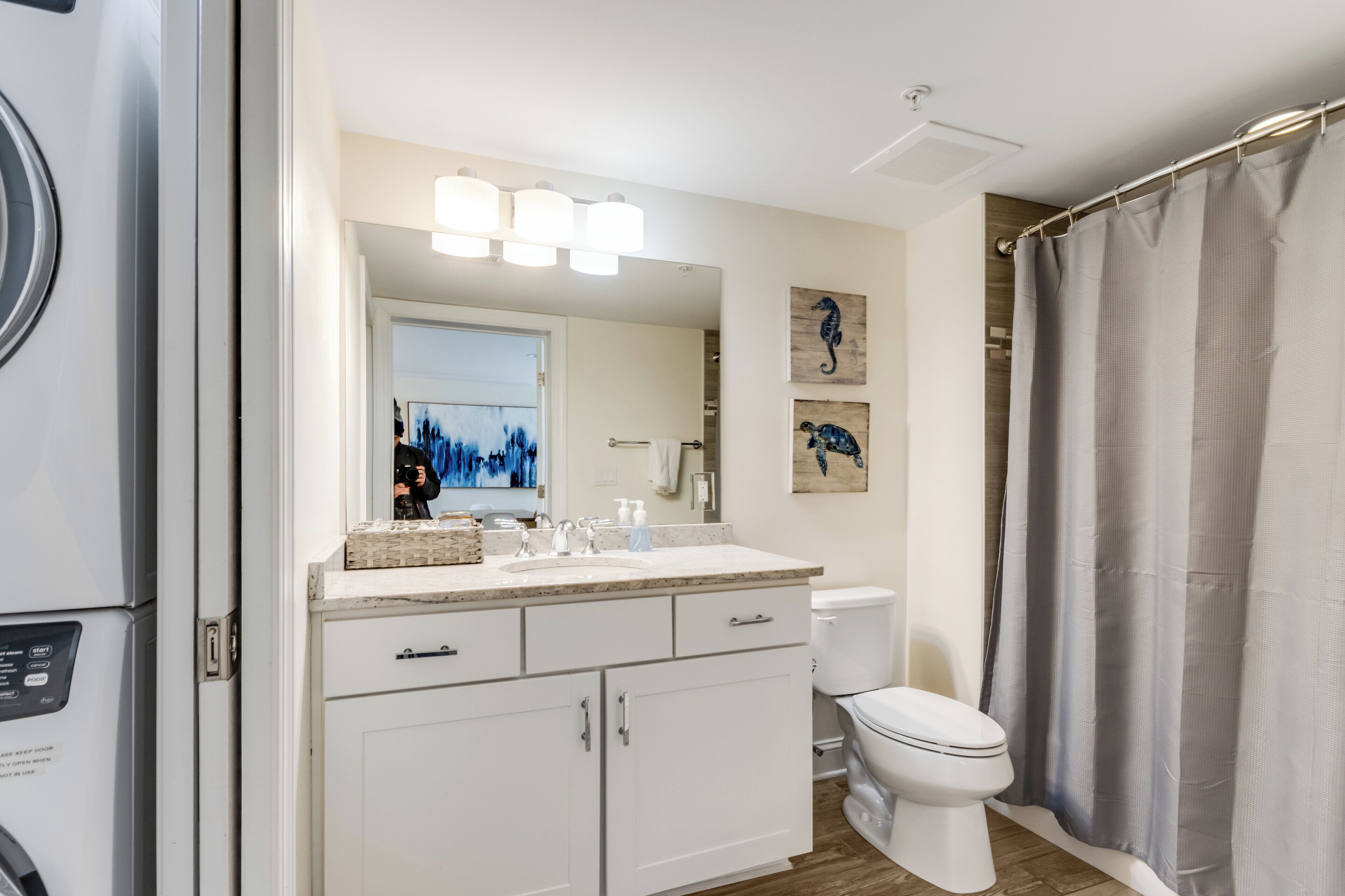 A modern bathroom with a white sink, mirror, and toilet. There are two framed artworks on the wall, a shower curtain, and a visible laundry machine.