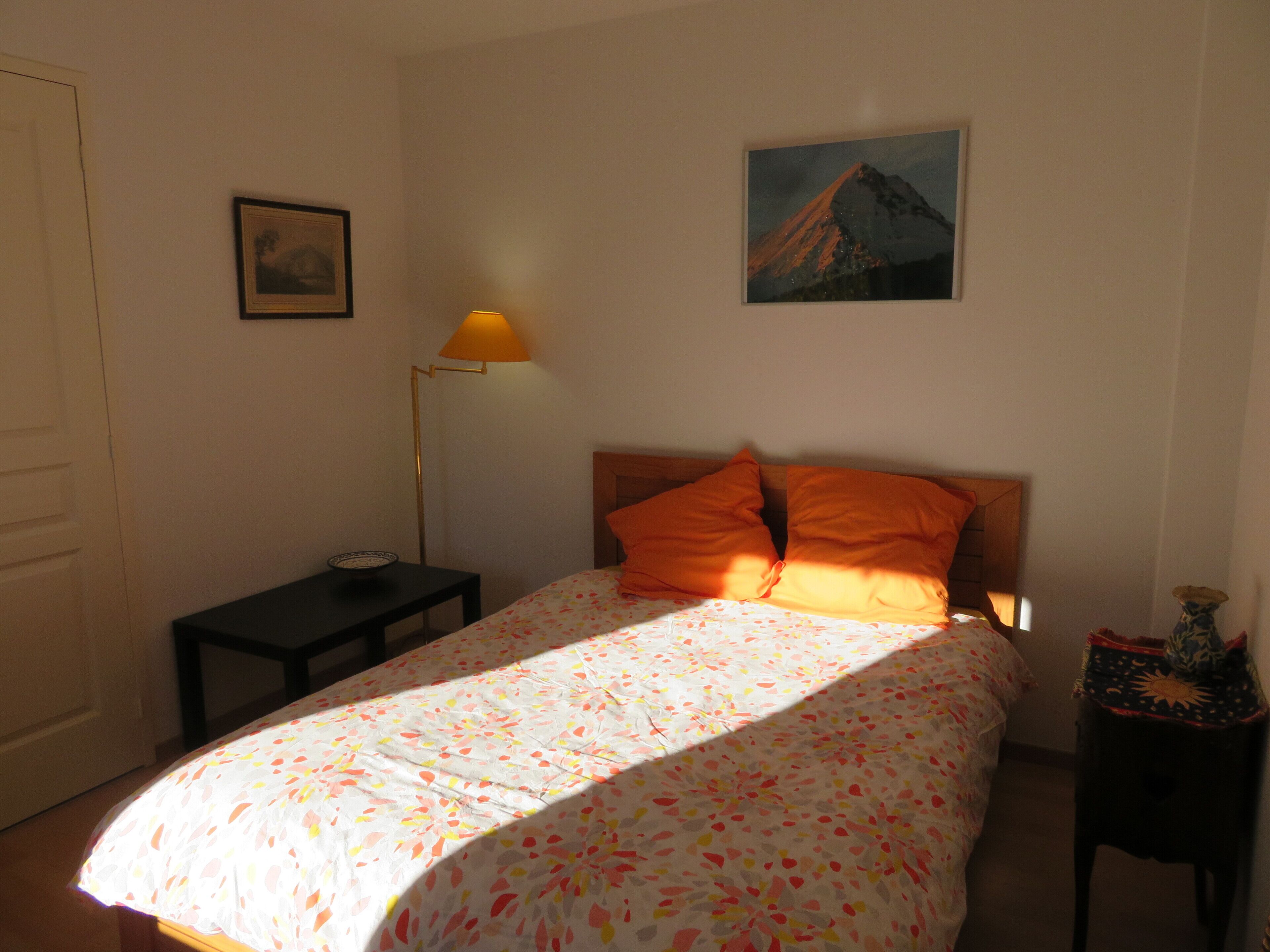 Une chambre à coucher avec un lit, une table de chevet, une lampe, un tableau encadré au mur et un vase sur la table de chevet.