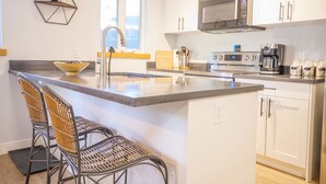 Una cocina moderna con armarios blancos, microondas de acero inoxidable, cafetera y barra de desayuno con dos sillas de mimbre.