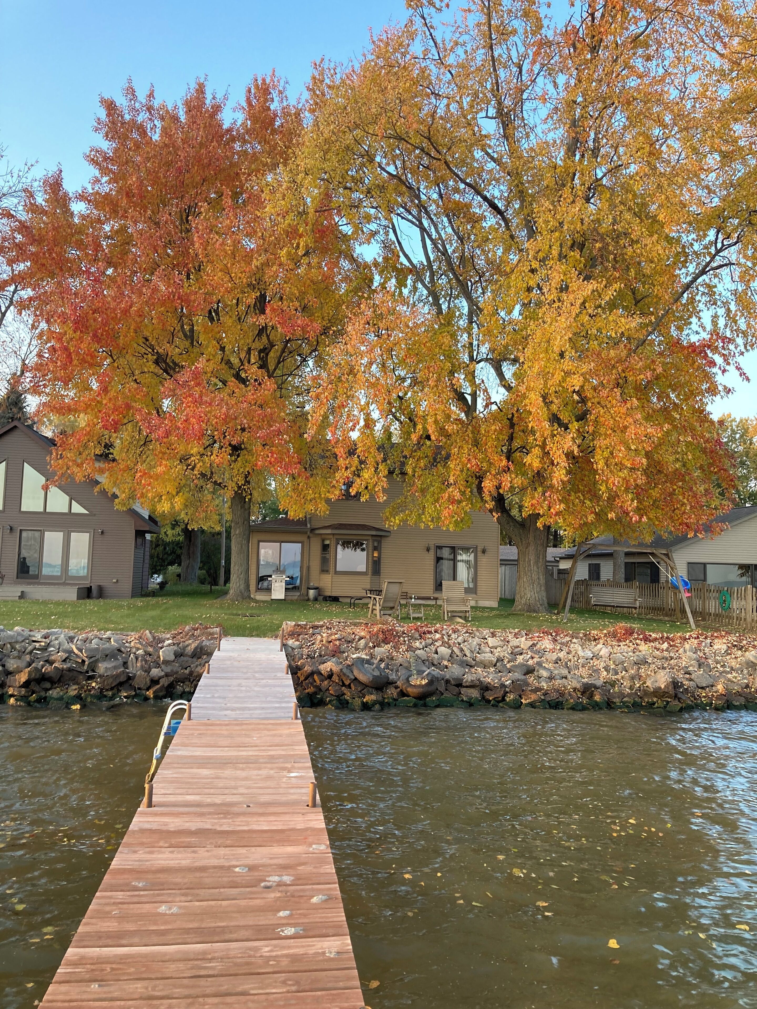 A wooden dock extending into a calm body of water, surrounded by autumn-colored trees and a row of houses with a stone wall and a grassy lawn.