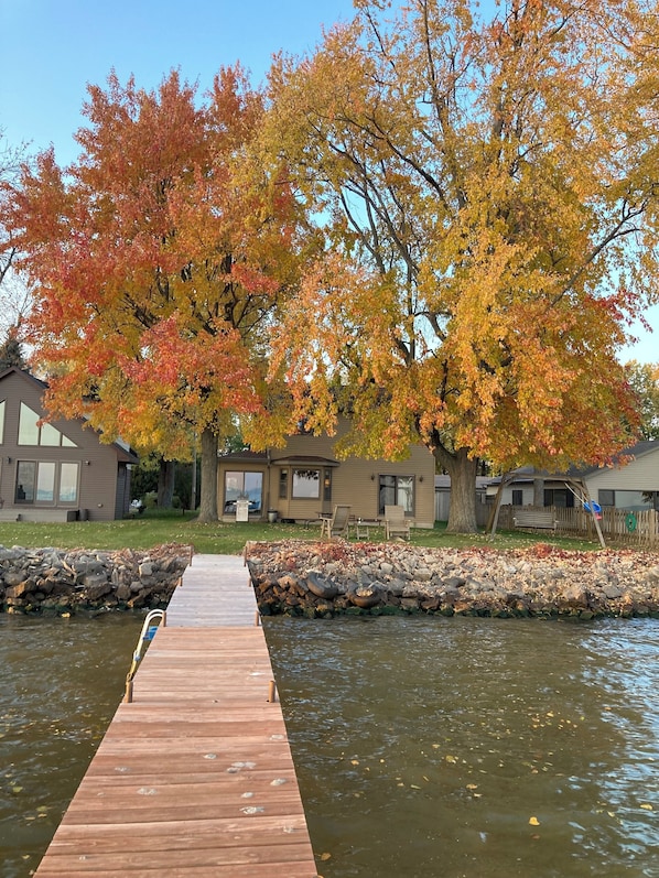 Une jetée en bois s’étendant sur une étendue d’eau calme, entourée d’arbres aux couleurs automnales et d’une rangée de maisons avec un mur de pierre et une pelouse verdoyante.