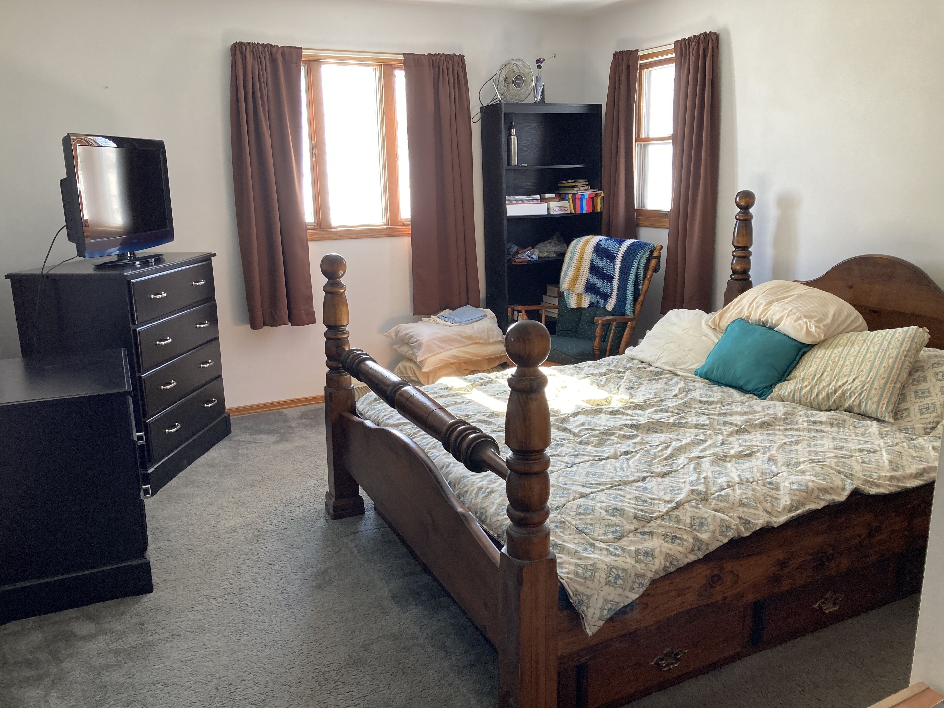 Una camera da letto con un letto di legno, una cassettiera, un televisore su un mobile, una libreria e una finestra con tende.
