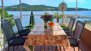 A covered outdoor dining area with a table set for two, chairs, a floral tablecloth, a vase with flowers, a bowl of oranges, and a bottle of wine.