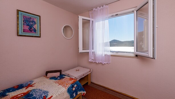 A bedroom with a single bed, a window with a curtain, a framed picture on the wall, and a small round mirror.