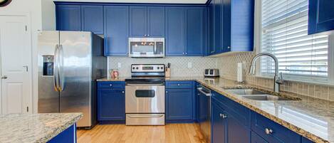 A modern kitchen with blue cabinets, stainless steel appliances, a granite countertop, and a window with blinds.