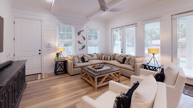 Un salón amplio con un sofá seccional grande, una mesa de centro y un ventilador de techo. Hay ventanas grandes con persianas, una chimenea y un televisor montado en la pared.