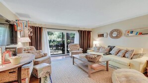 Una sala de estar con un sofá, sillones, una mesa de centro y un televisor. Hay una puerta corrediza de vidrio que da a un área exterior.