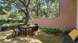 Un patio exterior con mesa y sillas, rodeado de plantas en macetas y un árbol grande.