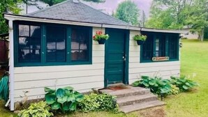 A small, single-story house with a green door and windows, surrounded by a grassy yard and large green plants. - Kawartha Lakes Cottage (Lakefield)