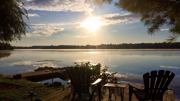 A lakeside deck with Adirondack chairs and a table, overlooking a calm lake with a sunset in the background. - Kawartha Lakes Cottage (Lakefield)
