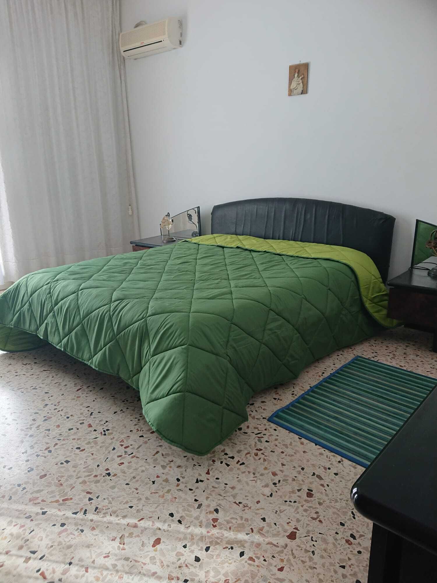 A bedroom with a green quilted bedspread, a black headboard, a small bedside table with a lamp, and a wall-mounted air conditioner.