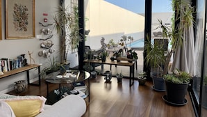 A modern living room with a large glass door leading to a balcony, wooden flooring, a coffee table with books and decorative items, and various potted plants. - Praia de Santos, Exclusive Villa, Sea-sided with swimming pool, Azores Islands (São Roque)