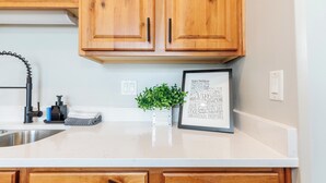 A kitchen with wooden cabinets, a white countertop, a sink, a faucet, a soap dispenser, a towel, a framed picture, and a small potted plant. - Beautiful, Sparkling Clean 3 BR, 2.5 BA Townhome!! (Providence)