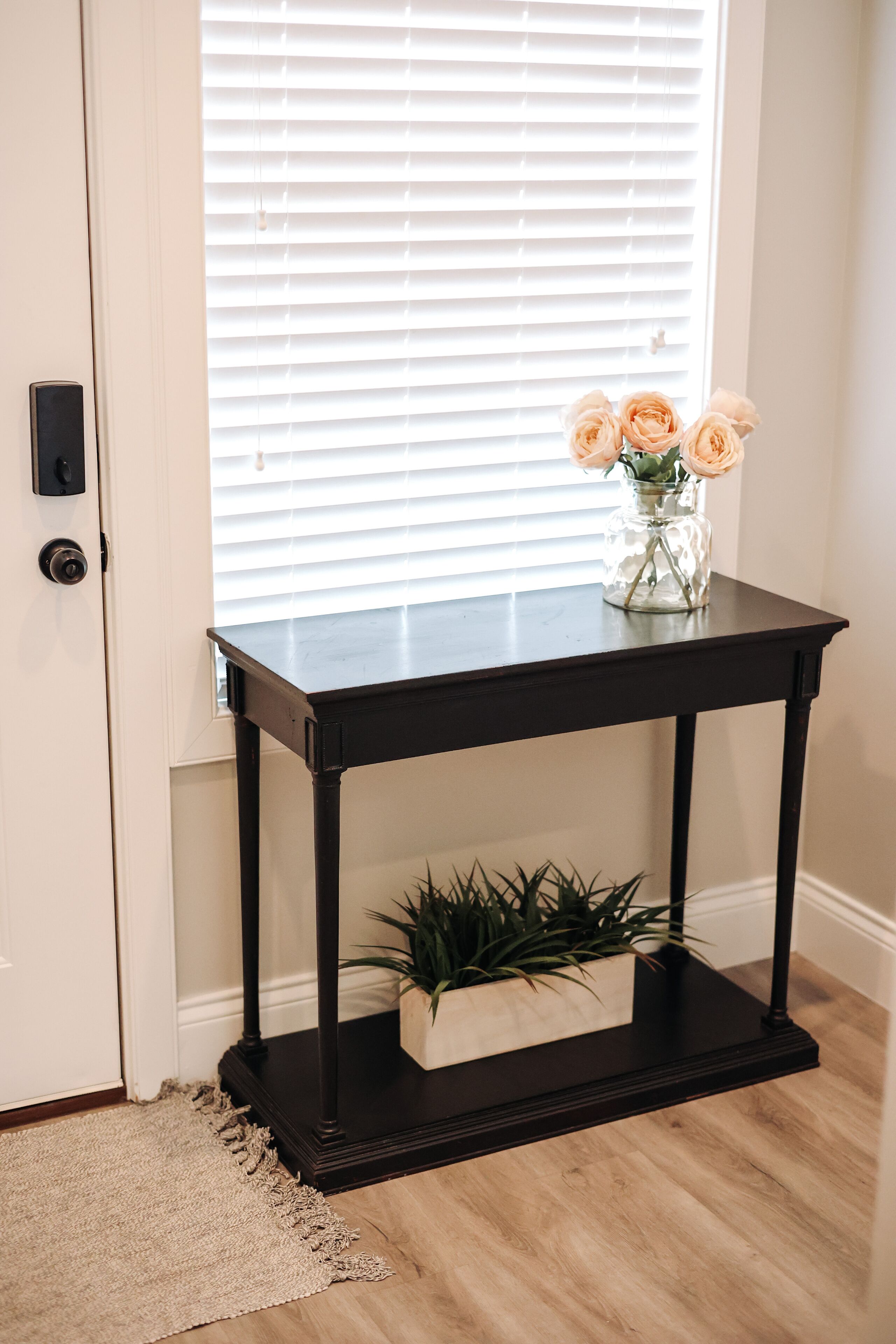 Une console en bois noir, sur laquelle sont posés un vase de fleurs et une plante, le tout sur un plancher en bois.