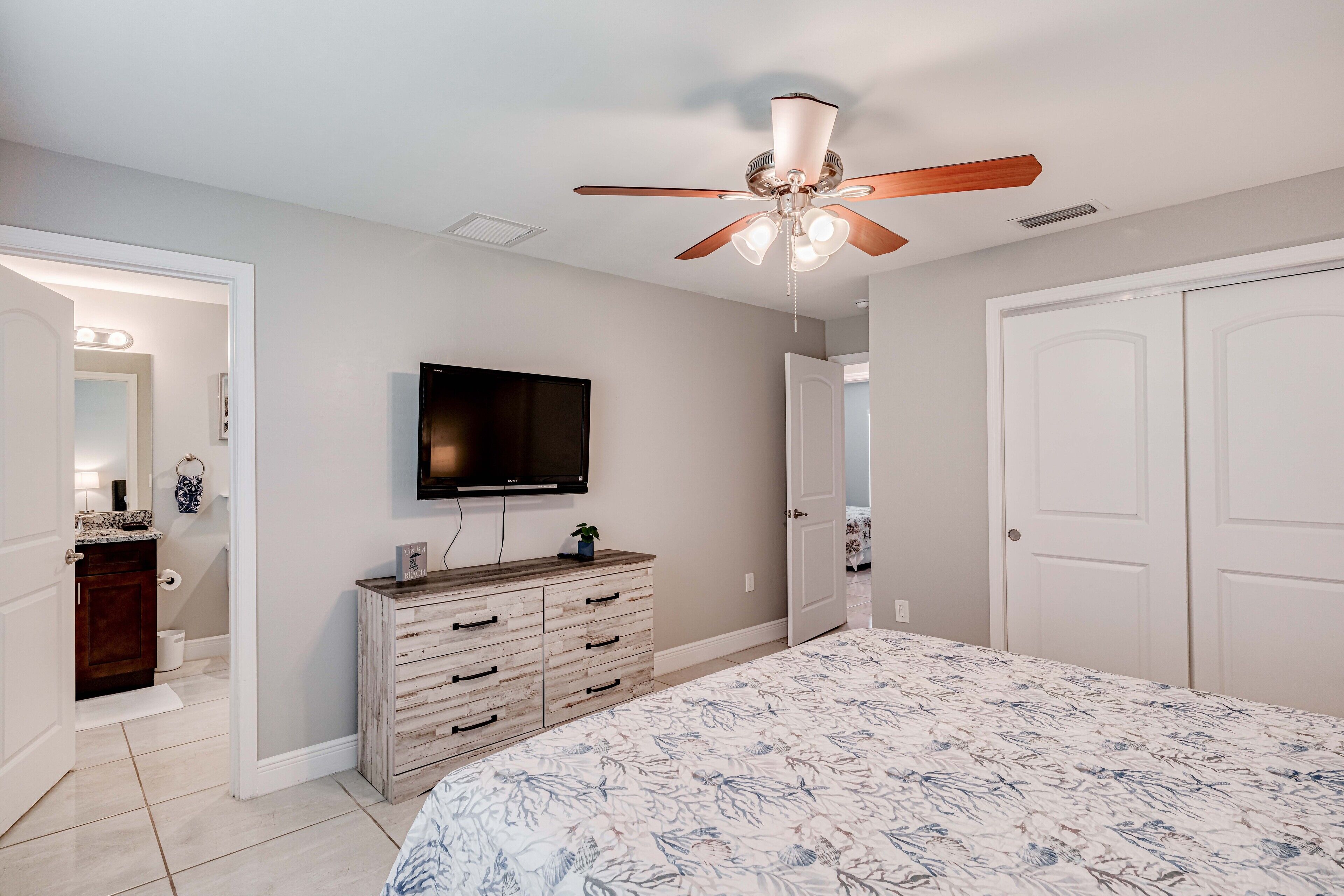A bedroom with a bed, a ceiling fan, a TV, a dresser, and a door leading to another room.