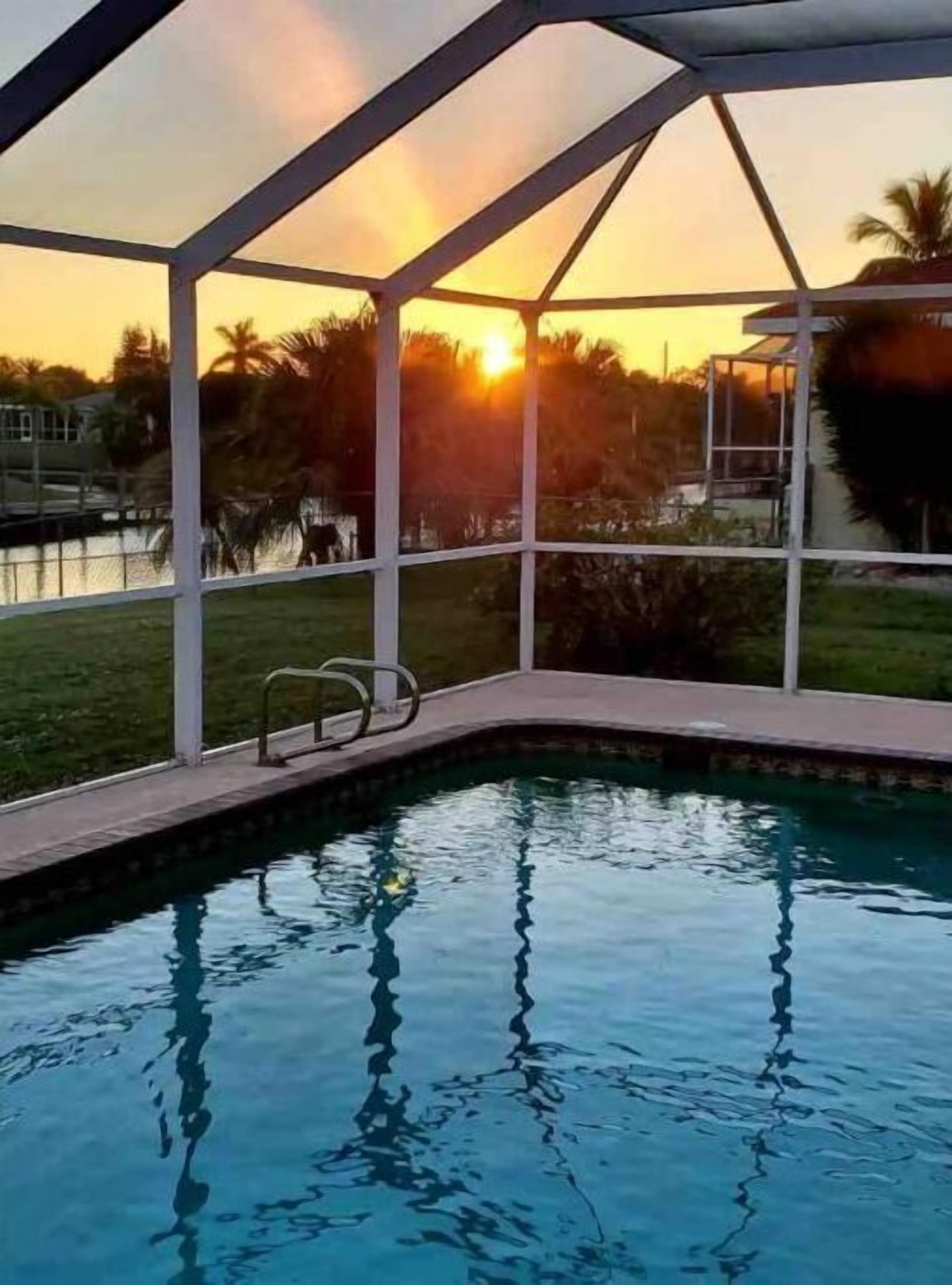Une piscine entourée d’une structure en verre, bordée d’une pelouse et de palmiers, avec le soleil couchant en arrière-plan.