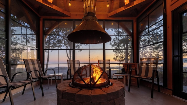 A cozy indoor seating area with a stone fire pit, large windows, and wooden beams. - Bois Blanc Island Lighthouse Home. Iconic Sunset Views of the Mackinac Bridge! (Bois Blanc Island)