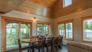 A dining area with a wooden table and chairs, large windows with a view of trees and water, and a wooden ceiling. - Bois Blanc Island Lighthouse Home. Iconic Sunset Views of the Mackinac Bridge! (Bois Blanc Island)