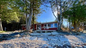 A red cabin with a stone foundation, surrounded by trees and a gravel path. - Waterfront gem, sandy beach, modern kitchen, large great room, free firewood (Sturgeon Bay)